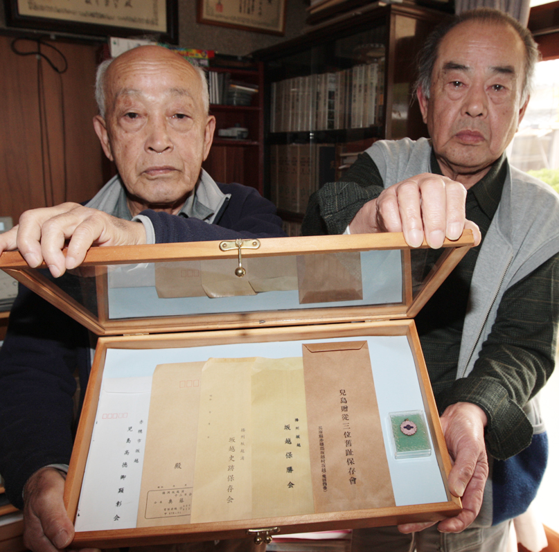 封筒に印刷された団体名で顕彰会の変遷を振り返る春野さん(右)と若松さん。ケースの右にあるのは明治40年制作の発足記念バッジ